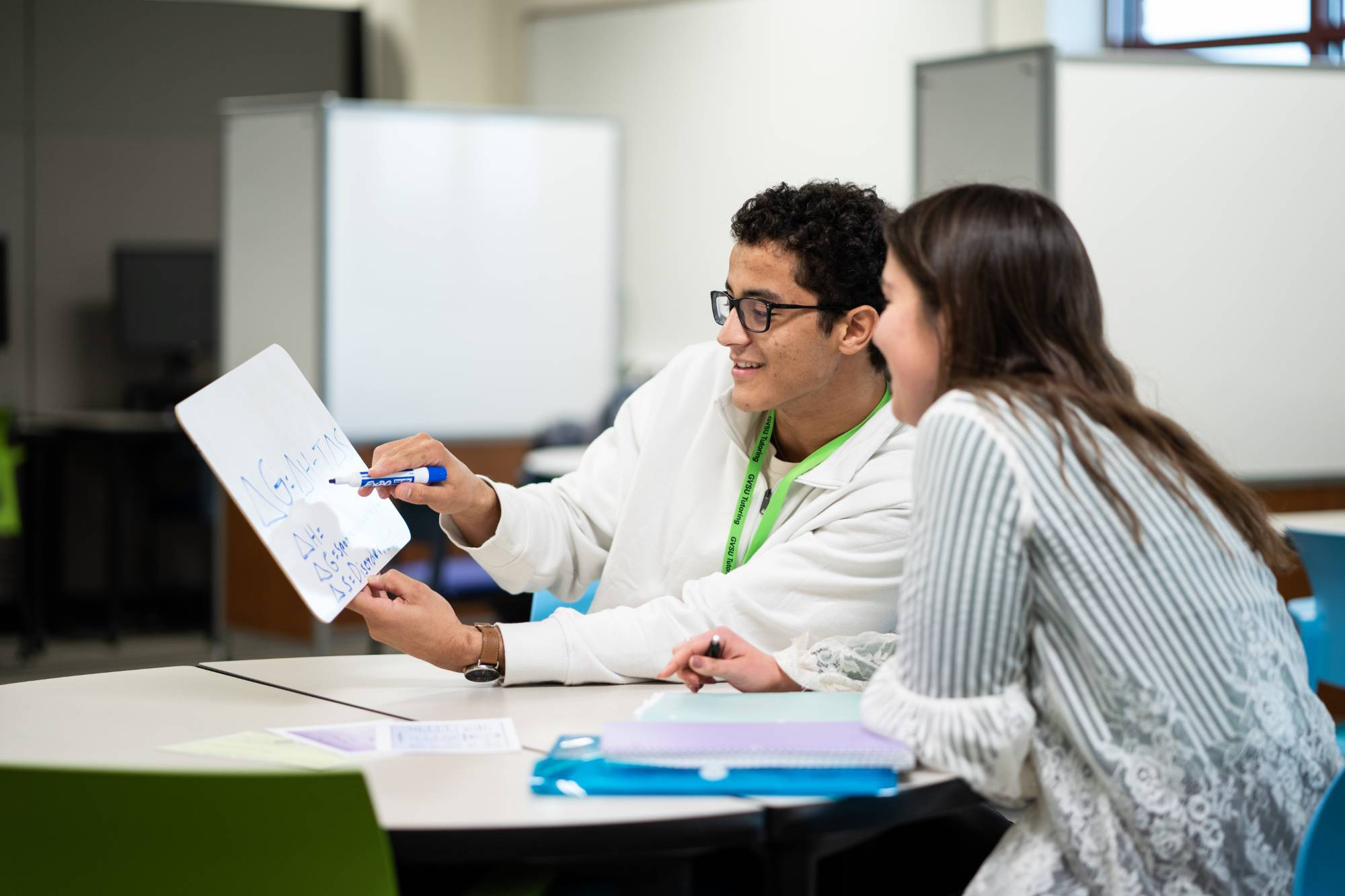 Tutor wearing glasses and green lanyard explains handwritten notes on paper to student during one-on-one session at table with study materials in tutoring center with cubicle workspace visible in background.